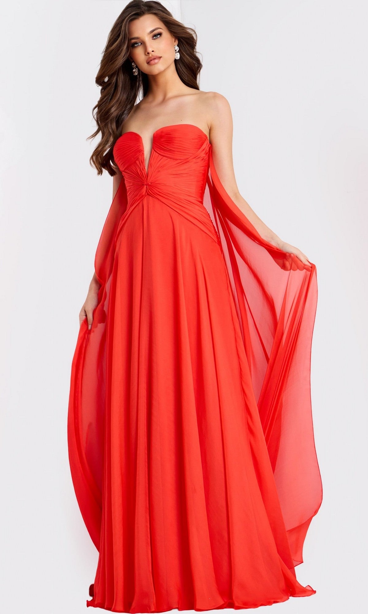 A woman poses against a plain light background wearing the Jovani 45269 long prom dress in orange, featuring a strapless chiffon design with a fitted bodice, pleated A-line skirt, and flowing sheer cape-like panels.